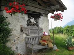 Teddybär auf einer Bank vor einem mit Blumen geschmückten Häuschen